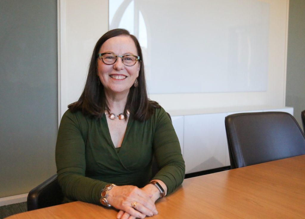 Woman in glasses and sweater sitting at desk, smiling.