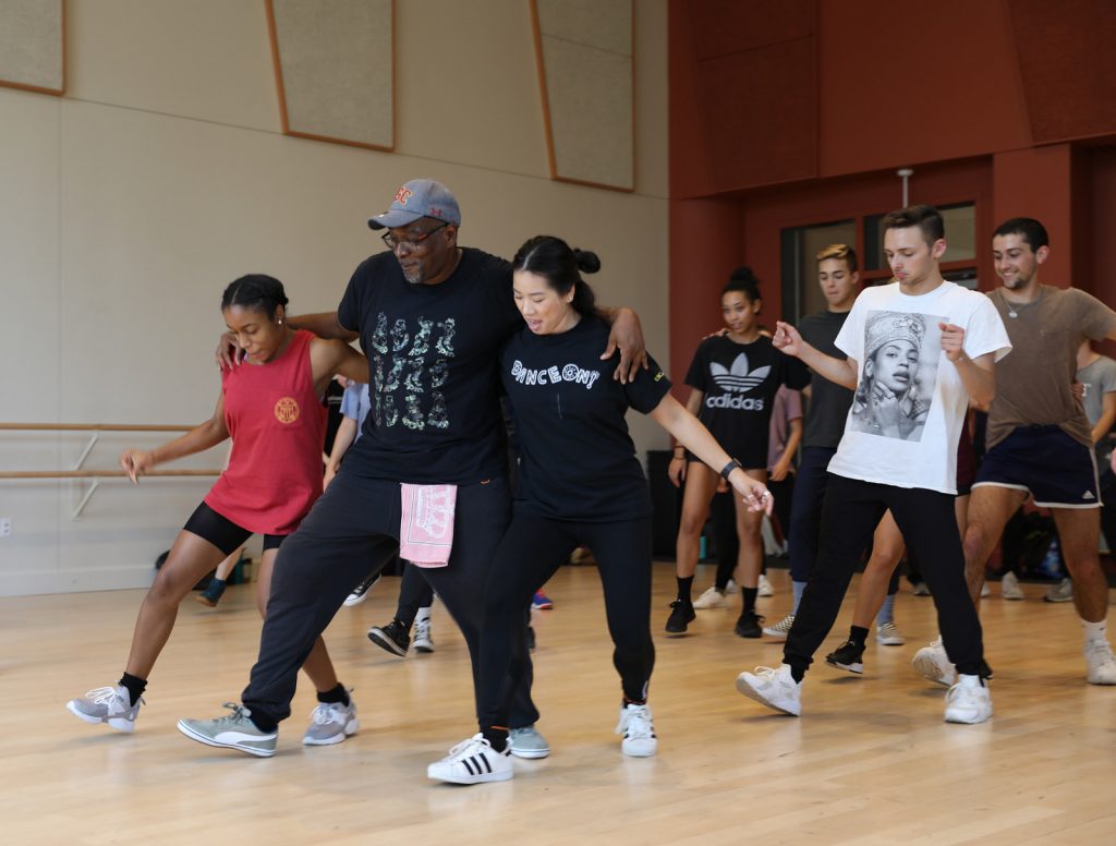 Teacher with his arms around two students as he teaches them dance steps in a studio.