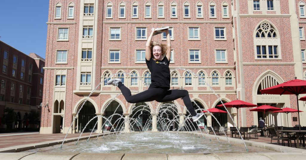 Girl jumps with her arms in the air in front of a water feature in the USC Village