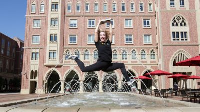 Girl jumps with her arms in the air in front of a water feature in the USC Village