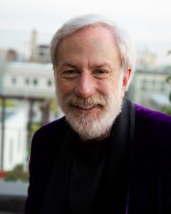 man with white hair and facial hair smiling and wearing a purple blazer