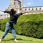 barefoot dancer reaching arms out wearing gray flannel shirt and blue jeans in front of green bushes