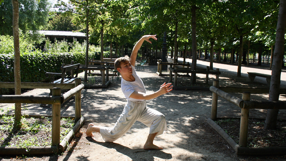 A boy dances in a park lined with trees and brown fences