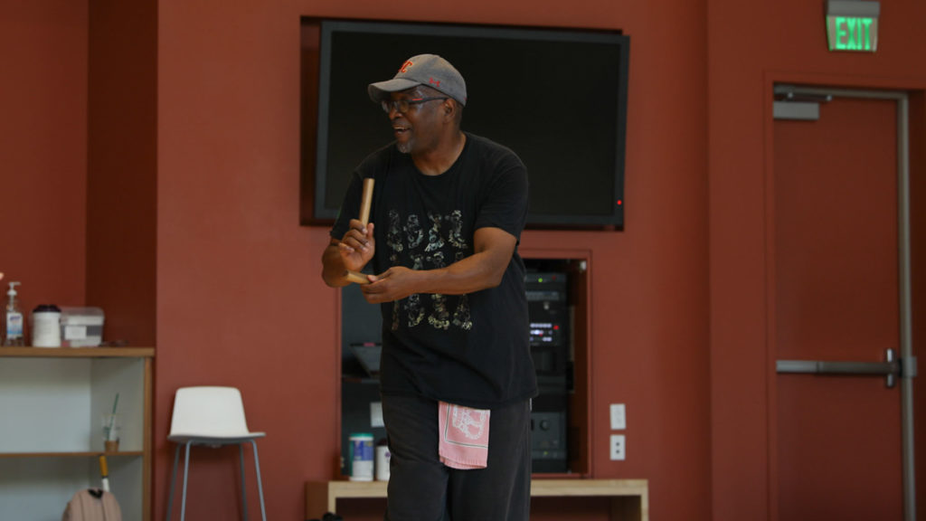 Moncell, dressed in black and a grey baseball hat, hits two sticks together in the dance studio