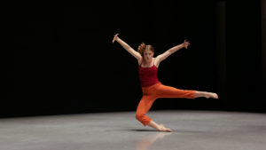 India, in a red tank top and orange capris, dances alone on a dark stage