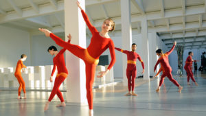 Dancers in red, orange, and yellow unitards strike geometrical poses throughout a white, industrial space