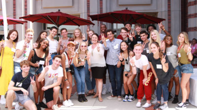 A group of USC Kaufman students pose around Deans Cutietta and Gates, all holding up a hand with their fingers in a 