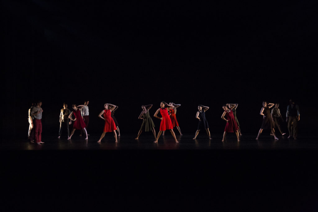 A large group of dancers in dresses strike the same angular pose on a dark stage 