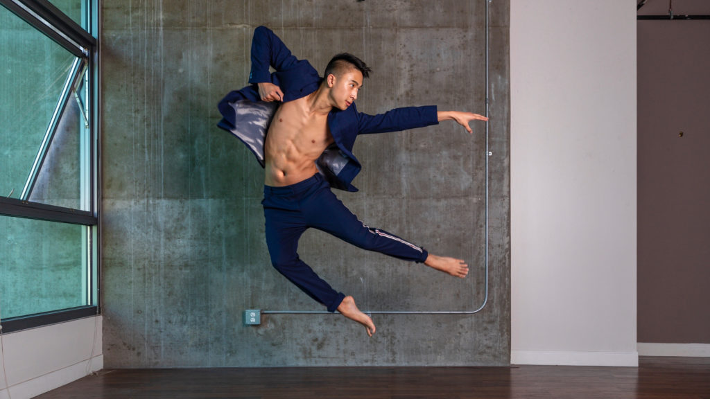 A dancer in blue jumps in front of a grey concrete wall