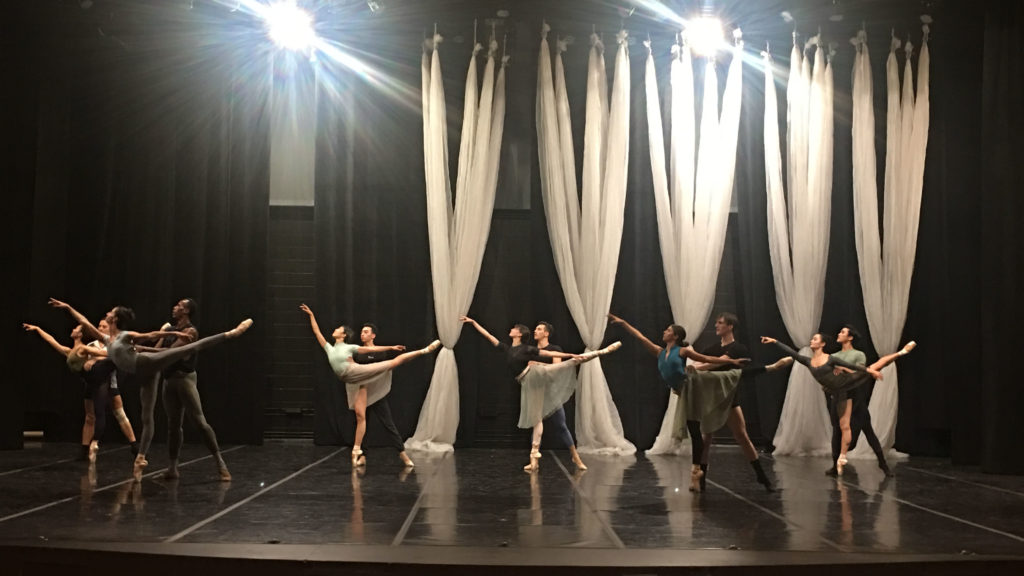 6 dancing couples pose in a partnered arabesque on a rehearsal stage