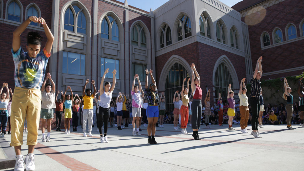 A large group of dancers outside of the Glorya Kaufman International Dance Center balance with their arms extended above their heads
