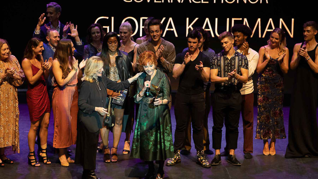 A woman in a green gown is surrounded on stage as she hold an award and speaks into the microphone