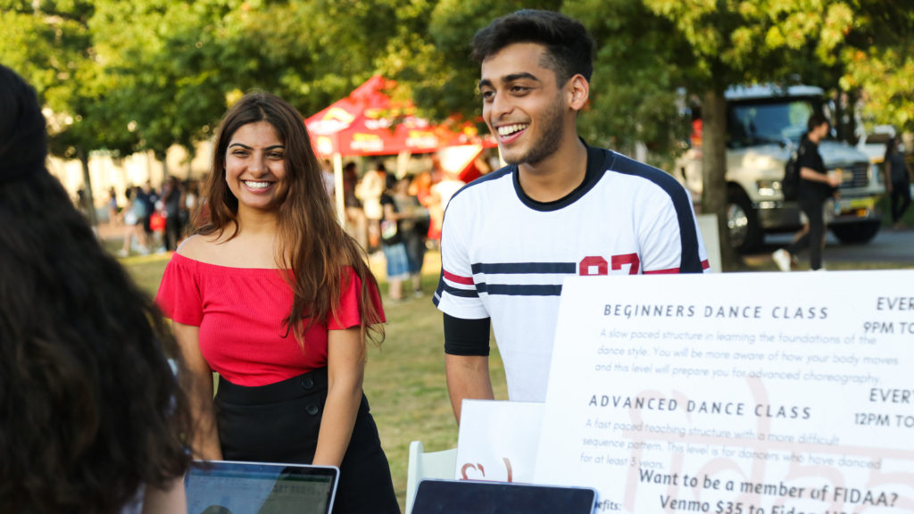 Two people at a booth smile as they talk to a passerby