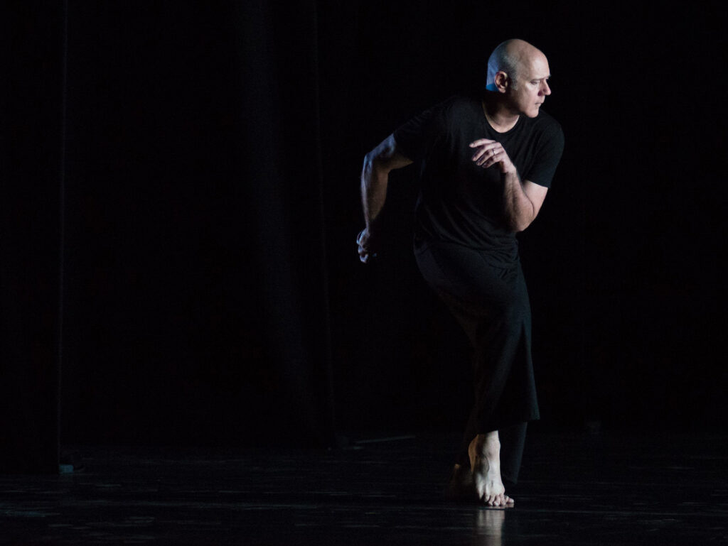 Doug Varone wearing all black dancing barefoot in front of a black background