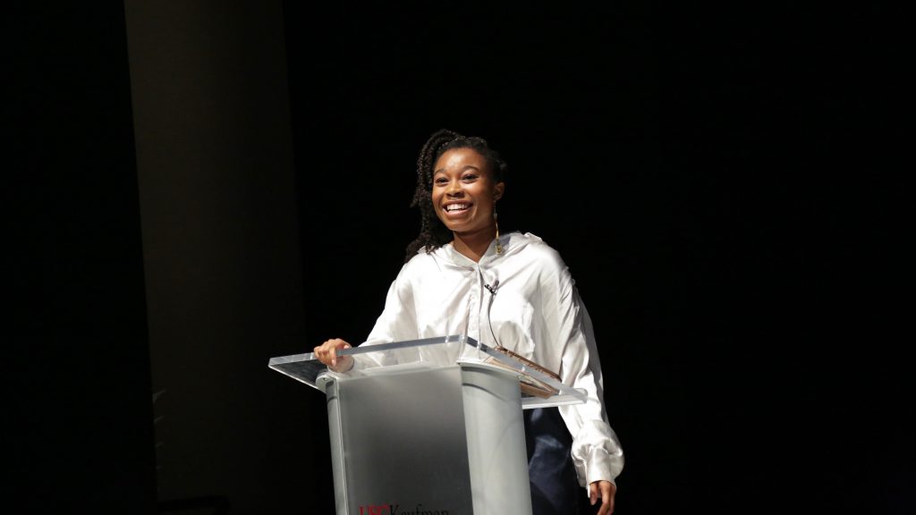 A woman wears a white windbreaker jacket and stands behind a podium giving a presentation