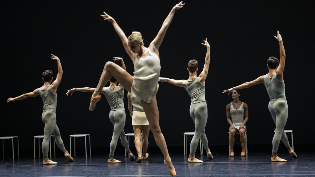 A dancer in the foreground jumps in an exaggerated side attitude, while a row of dancers pose behind her in an exaggerated fourth position