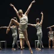 A dancer in the foreground jumps in an exaggerated side attitude, while a row of dancers pose behind her in an exaggerated fourth position