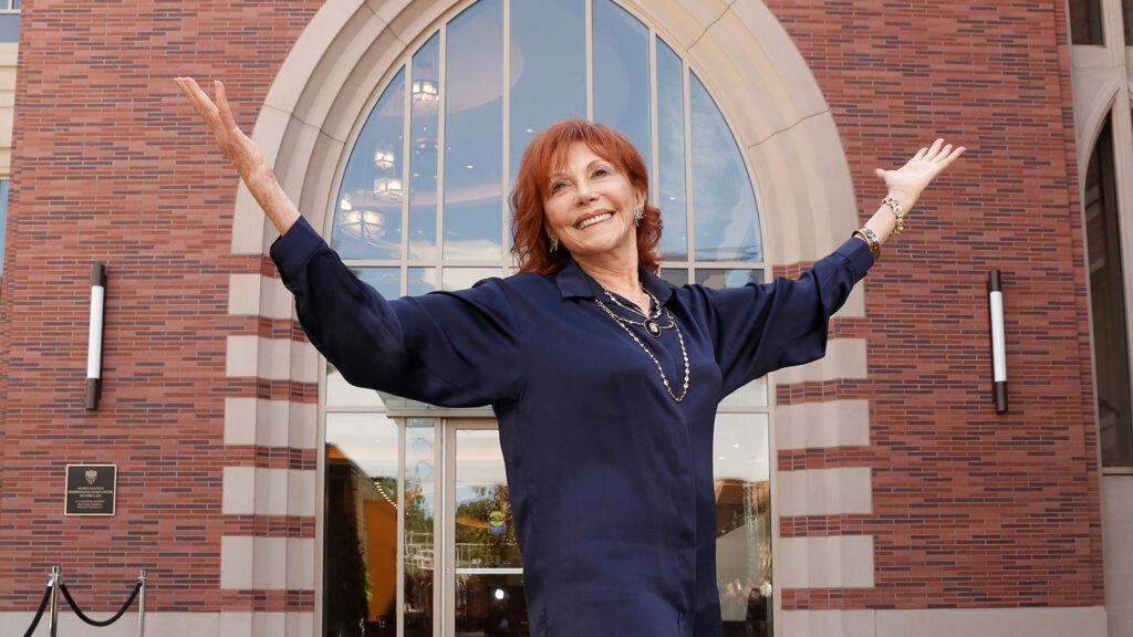Glorya Kaufman raising her hands and looking up in front of brick building