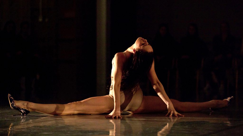 A woman in a beige leotard sits in the middle splits on stage.