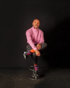 A man in a pink turtleneck sweater sits cross-legged on a stool.