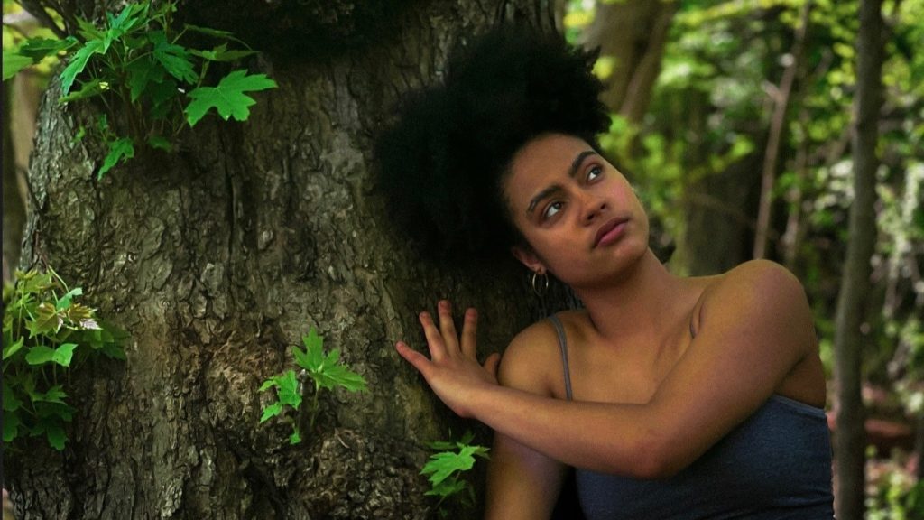 A woman wearing a gray tank top leans against a brown tree trunk with bright green leaves.