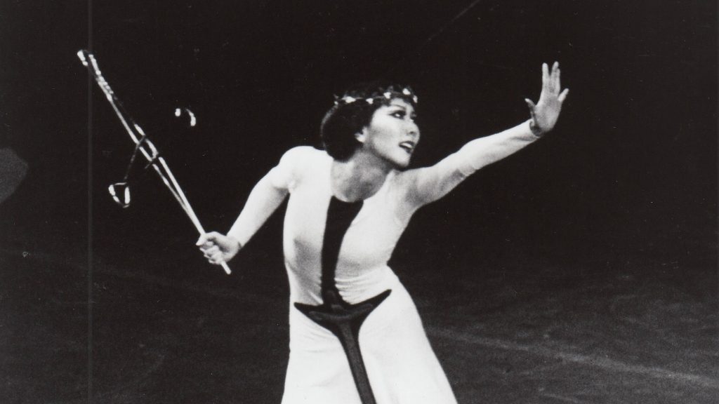 A woman wearing a black and white dress stands on stage and reaches out her hand while she looks beyond it.