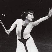 A woman wearing a black and white dress stands on stage and reaches out her hand while she looks beyond it.