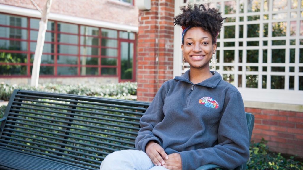 A woman sits on a green bench in front of a brick trellis. She is wearing a gray sweatshirt with the logo of Change the Stigma, which is shaped like a brain.