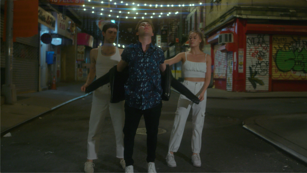 Two men and one woman stand in a triangle formation on a city street. The two dancers wearing white help the central male dancer take off his black leather jacket.