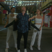 Two men and one woman stand in a triangle formation on a city street. The two dancers wearing white help the central male dancer take off his black leather jacket.