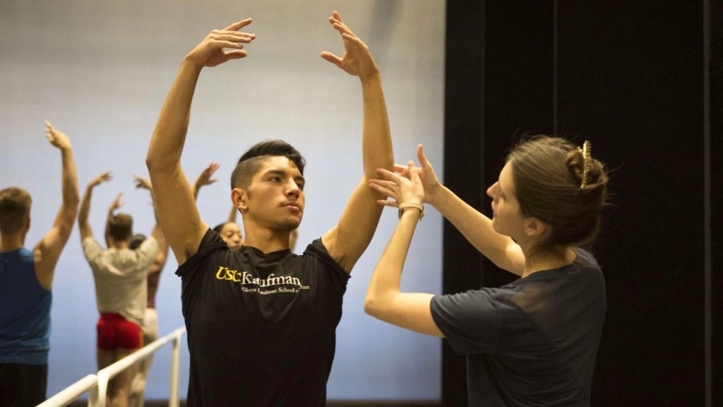 A woman wearing a black shirt guides a male ballet dancer's arms above his head.
