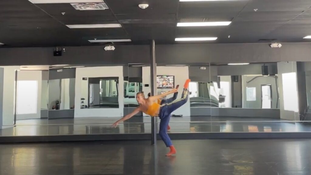 A dancer wearing an orange tank top and blue pants lifts their leg over their head and reaches their hand to the ground.