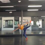 A dancer wearing an orange tank top and blue pants lifts their leg over their head and reaches their hand to the ground.