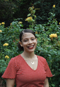 A woman in a red and white gingham shirt smiles in front of green leaves and yellow roses.