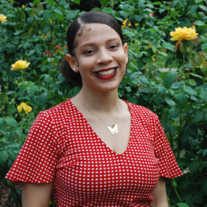 A woman in a red and white gingham shirt smiles in front of green leaves and yellow roses.