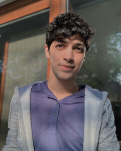 A man with dark hair wearing a purple button-up shirt and gray hoody smiles at the camera in front of green-tinted windows.