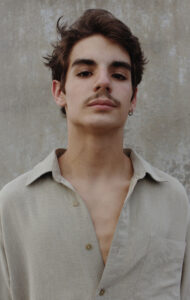 A man in a beige shirt stands in front of a beige wall and looks at the camera.