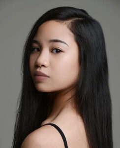 A woman with dark hair looks over her shoulder at the camera in front of a gray background.