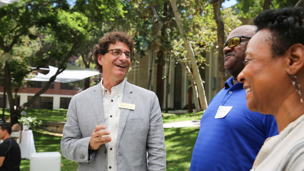 A man in a grey suit jacket and white button-down shirt speaks with a man in a blue polo shirt and a woman in a cream shirt.