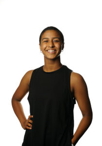 A person wearing a black tank top stands with her hand on her hip as they smile.