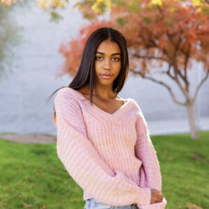 A woman with long, dark hair wearing a pink sweater and light blue jeans looks at the camera in front of grass and a tree with red-orange leaves.