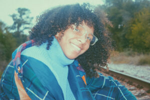 a person with brown curly hair and glasses on smiling over their shoulder with a turtle neck and jacket on