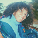 a person with brown curly hair and glasses on smiling over their shoulder with a turtle neck and jacket on