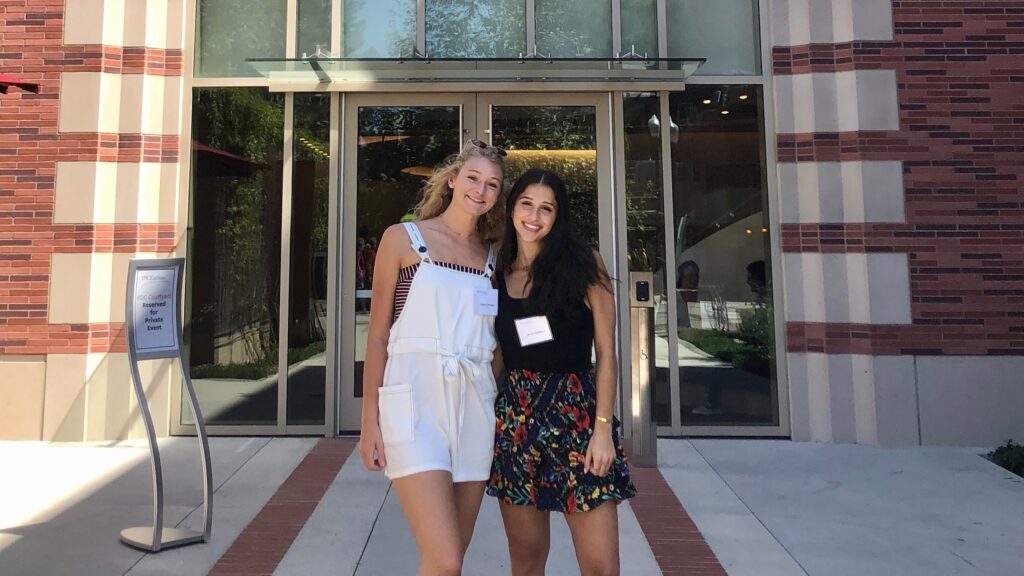 A woman with blonde hair wearing white overalls stands next to a woman in a black shirt and floral shorts in front of a large brick building.