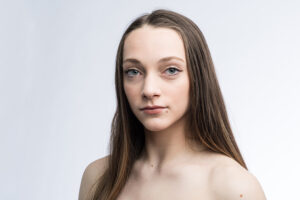 A woman with long, brown hair looks at the camera in front of a white background.