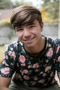 A man with light brown hair wearing a black t-shirt with pink roses smiles at the camera in front of trees and a wooden fence.