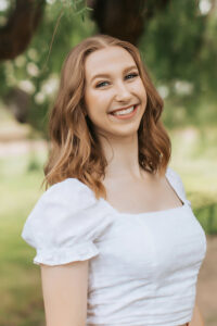 A woman with light brown hair wearing a white blouse smiles at the camera in front of green grass and trees.