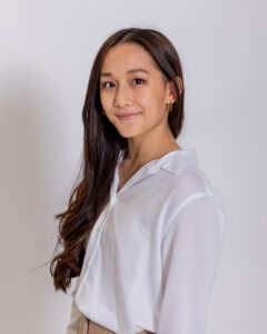 A woman with long, dark hair wearing a white blouse smiles at the camera in front of a white background.