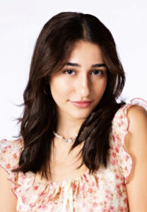 A woman with dark brown hair wearing a pink floral shirt smiles at the camera in front of a white background.