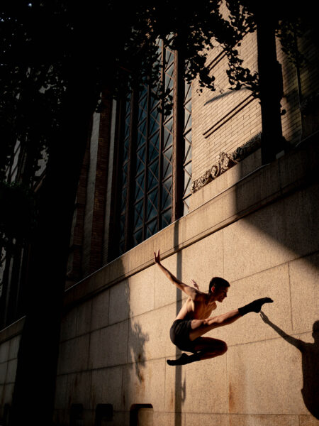 A man wearing dark shorts jumps in the air pointing his leg in front of him with his arms behind him in front of a concrete building with large windows.